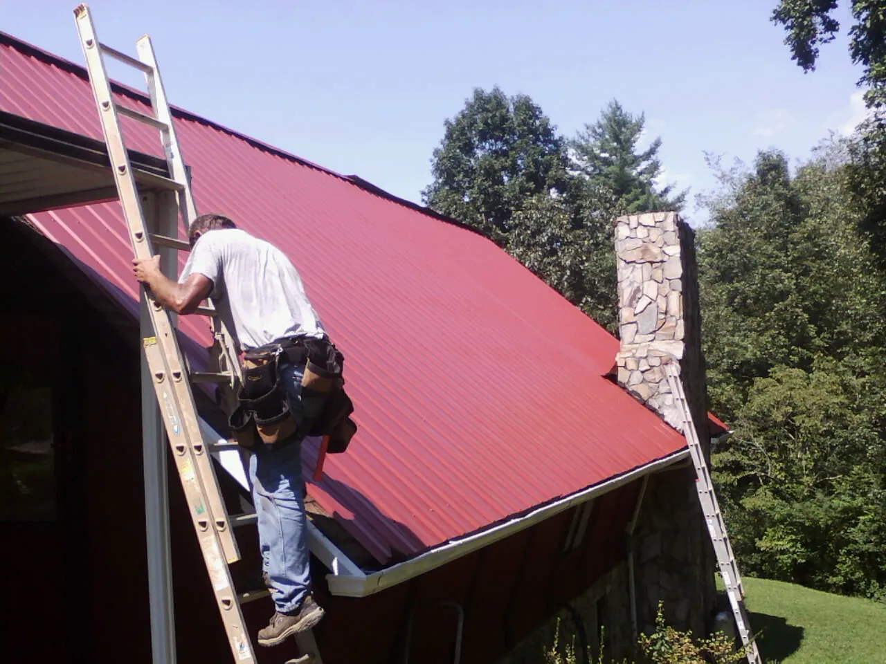 Roofer on a ladder beside a red metal roof and stone chimney — Soffit & Fascia Repair by Roofing Brothers in Seaview